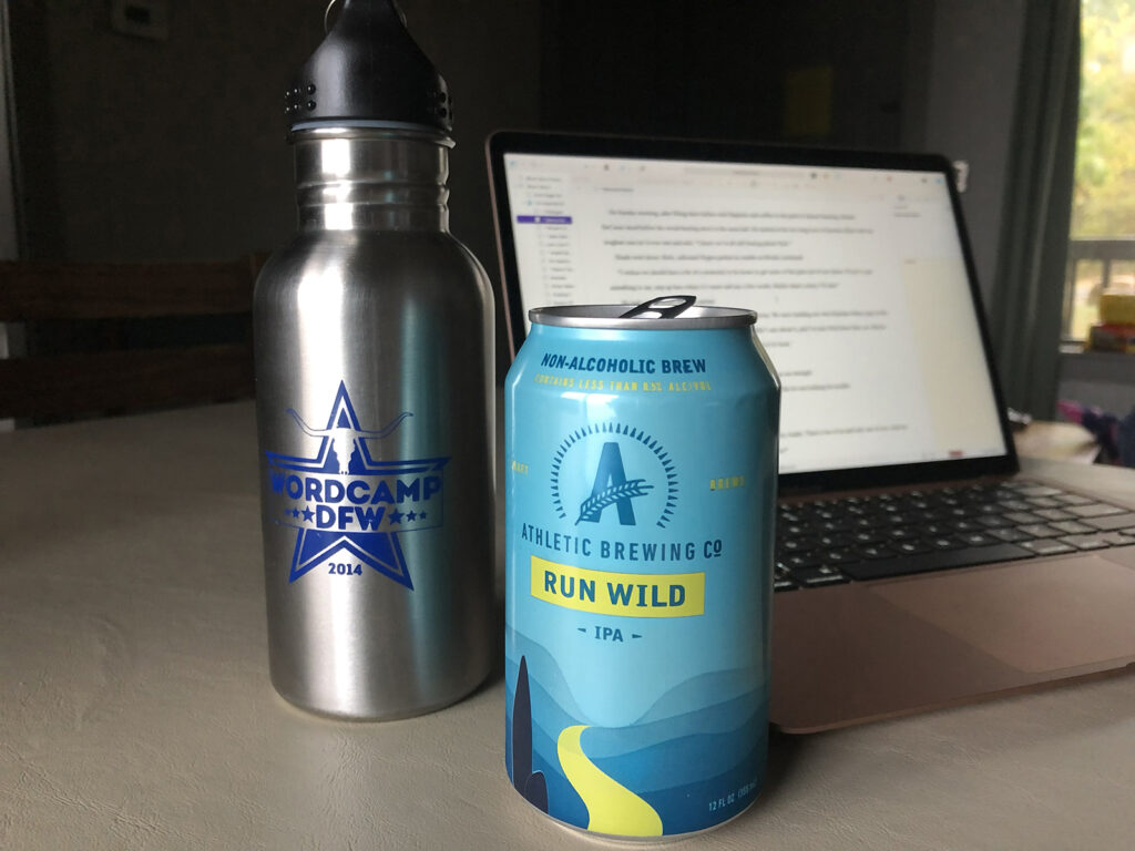 Water bottle reading WordCamp DFW 2014 next to a can of Athletic Brewing Company Run Wild non-alcoholic IPA. In the background, a laptop opened to a story in Scrivener.