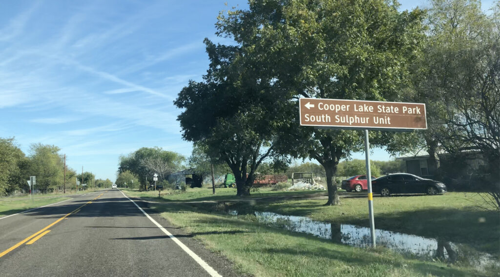 The road near Cooper Lake State Park (with a sign with an arrow pointing to the left and text: Cooper Lake State Park South Sulphur Unit.)