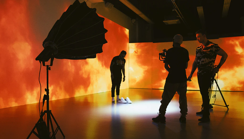 Videographers discuss a shot behind the scenes of a shoot in a warehouse. The subject stands waiting against a backdrop of orange clouds projected on the walls. A camera flash in a directional housing is set up in the left foreground.