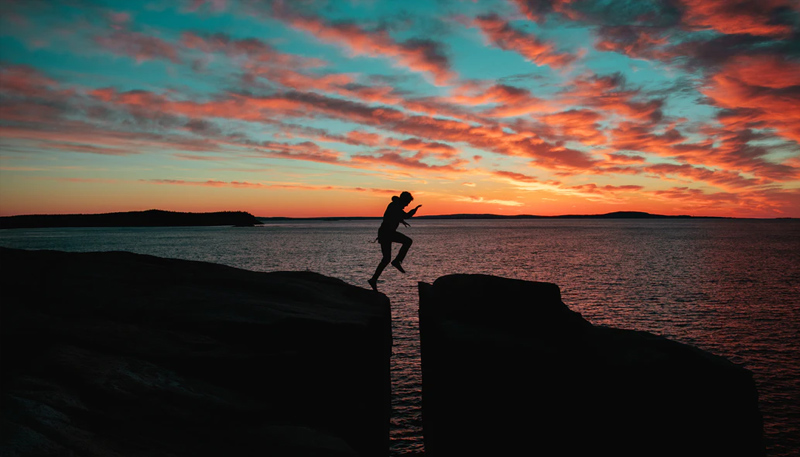 Silhouette of a person leaping over a gap between two rocks. Behind that, the ocean stretches to an orange sunset.