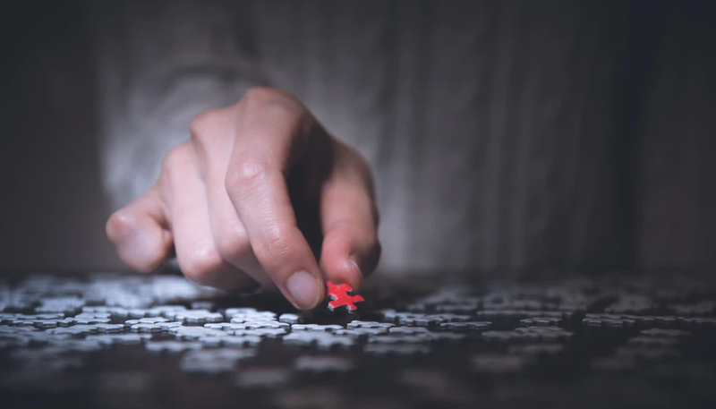 A hand places a red puzzle piece into a jumbled spread of white puzzle pieces.