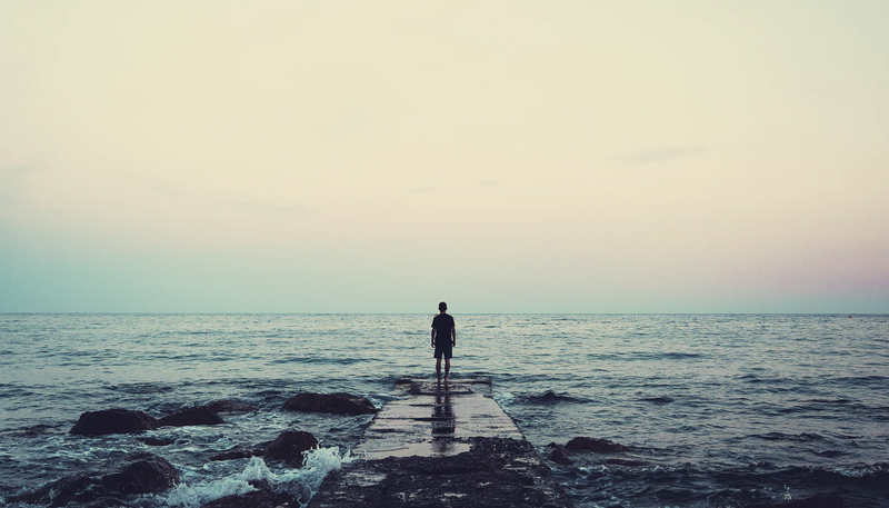 A person stands at the end of a concrete pier while staring at a roiling ocean.