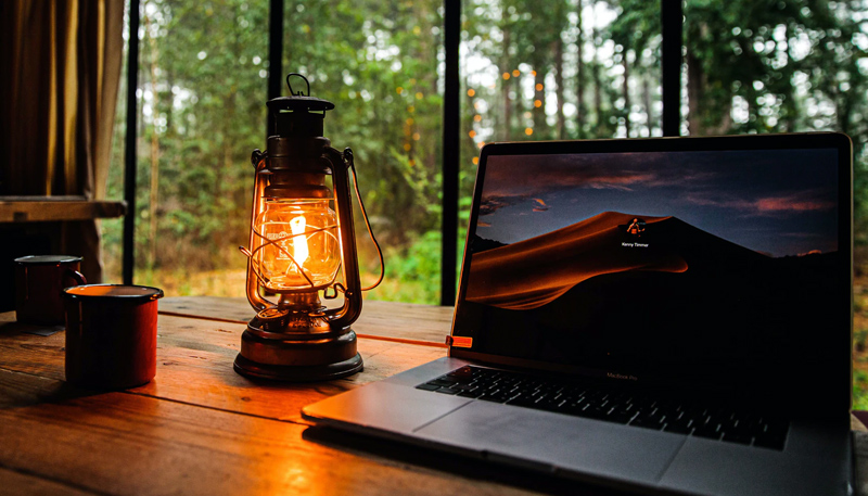 A wood-plank desk with (Left to Right) two metal camping mugs, a lit lantern, and a laptop computer.
Behind them, though a window, green trees at dusk...