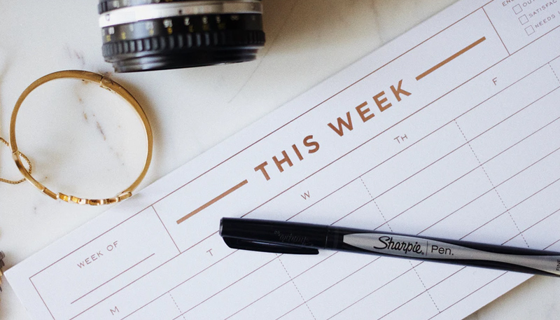 A Sharpie brand pen rests on top of a weekly calendar reading THIS WEEK across the top. Also in the photo, a bit of a camera lens.