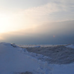 Person standing in the snow and ice as the sun shines on the horizon