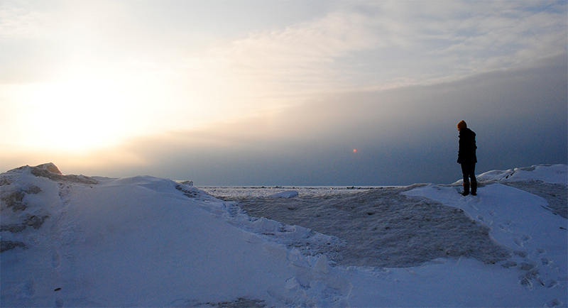 Person standing in the snow and ice as the sun shines on the horizon